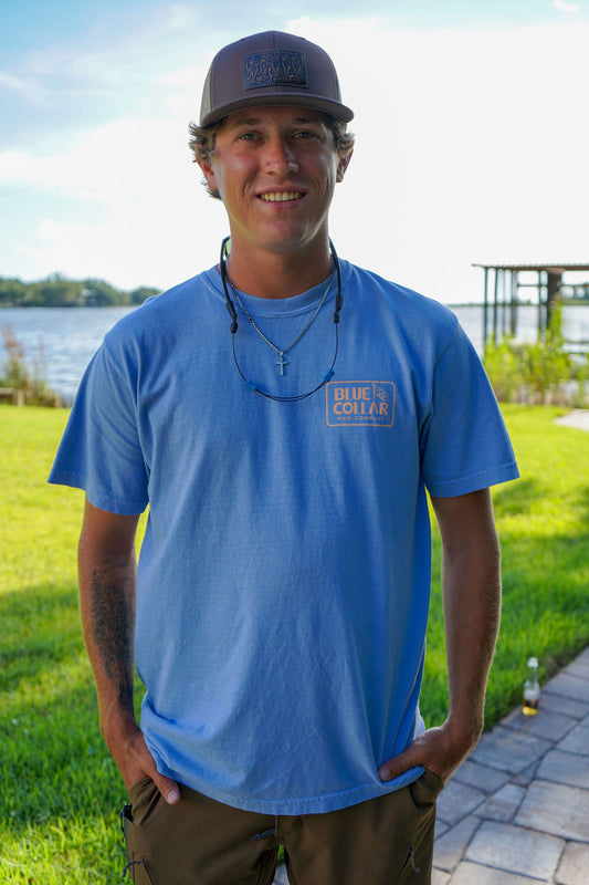 A smiling man wearing a light blue crewneck t-shirt with a 'Blue Collar' logo on the left chest, paired with a dark cap and brown pants, standing outdoors in front of a lake and green grass.