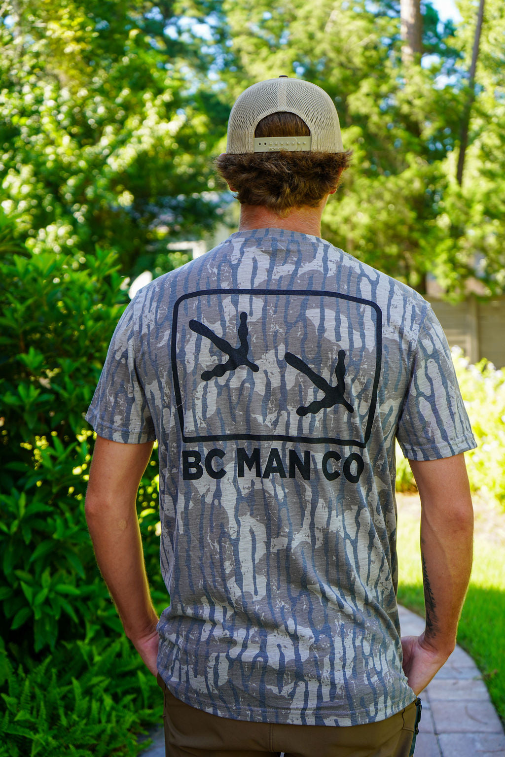 A man viewed from behind wearing a gray and blue camouflage t-shirt with a black graphic of two hands pointing outwards and the text 'BC MAN CO' printed below. He is wearing a beige baseball cap and standing outdoors on a paved path surrounded by green foliage.