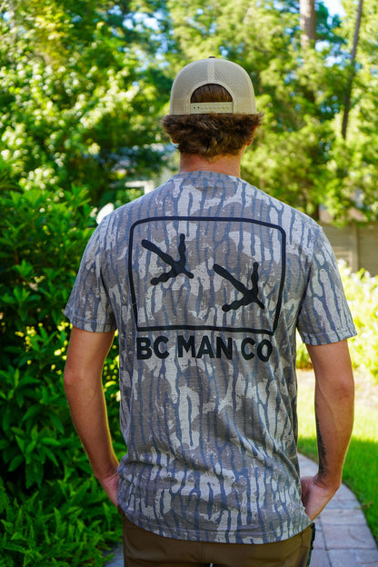 A man viewed from behind wearing a gray and blue camouflage t-shirt with a black graphic of two hands pointing outwards and the text 'BC MAN CO' printed below. He is wearing a beige baseball cap and standing outdoors on a paved path surrounded by green foliage.