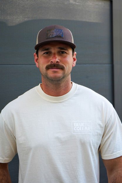 A man with a mustache wearing a brown baseball cap and a white t-shirt with a 'Blue Collar' logo on the chest, standing against a dark gray paneled wall.