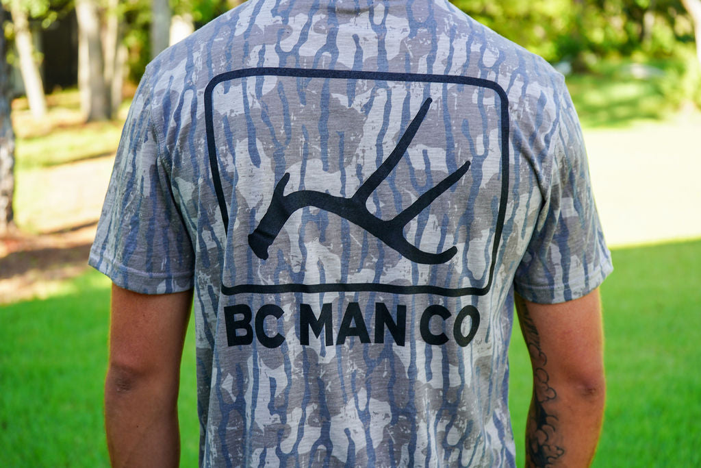 A man viewed from behind wearing a short-sleeve t-shirt with a blue, gray, and white camouflage pattern and a large black graphic of antlers with the text 'BC MAN CO' underneath, set against a blurred green park background.
