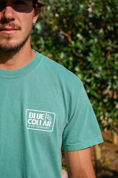 A man wearing a teal-green crewneck t-shirt with a white 'Blue Collar Man Company' logo printed on the left chest, set against a blurred green outdoor background.