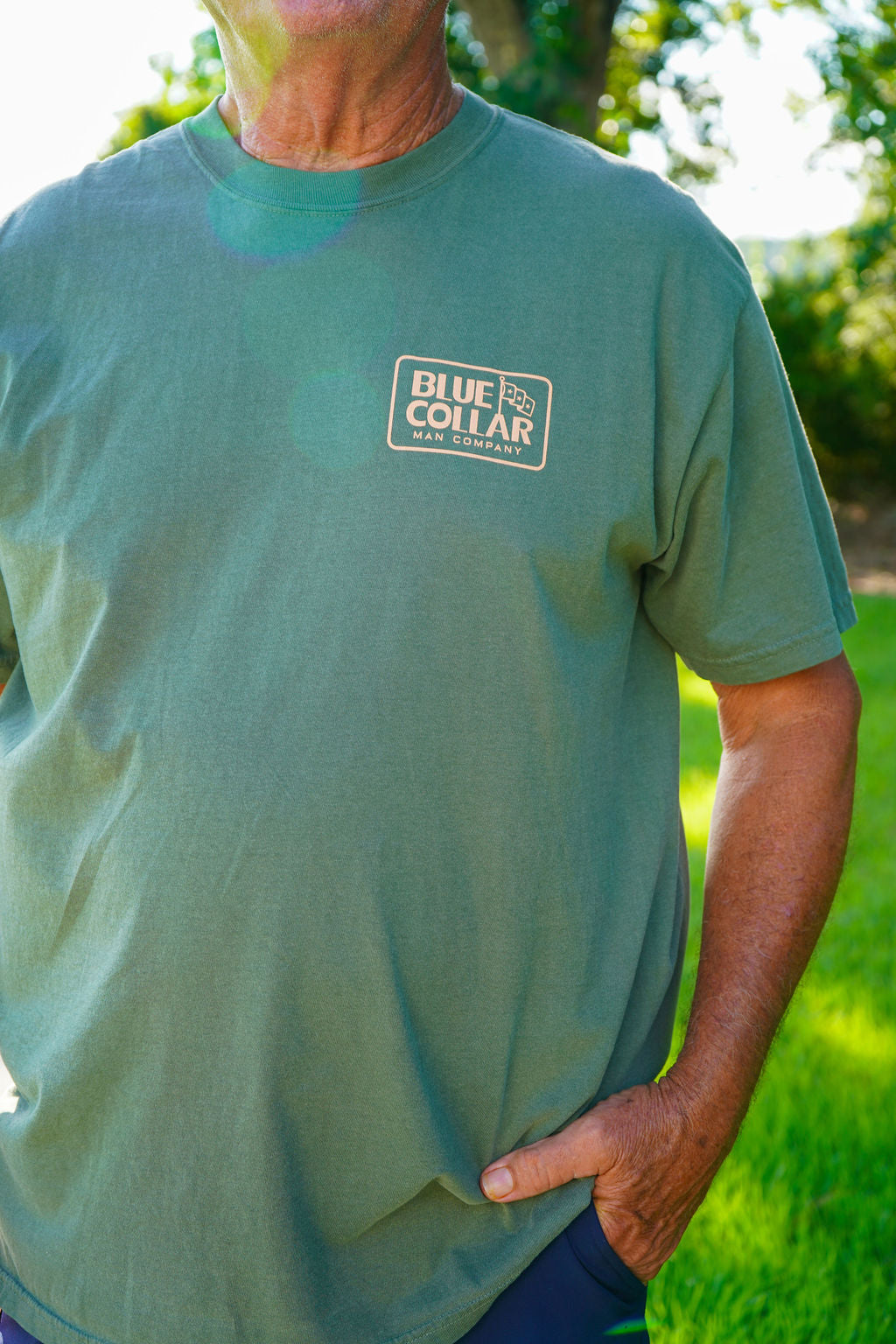 A man wearing a sage green crewneck t-shirt with a white 'Blue Collar Workwear' logo patch on the left chest, photographed outdoors in a sunny grassy area.