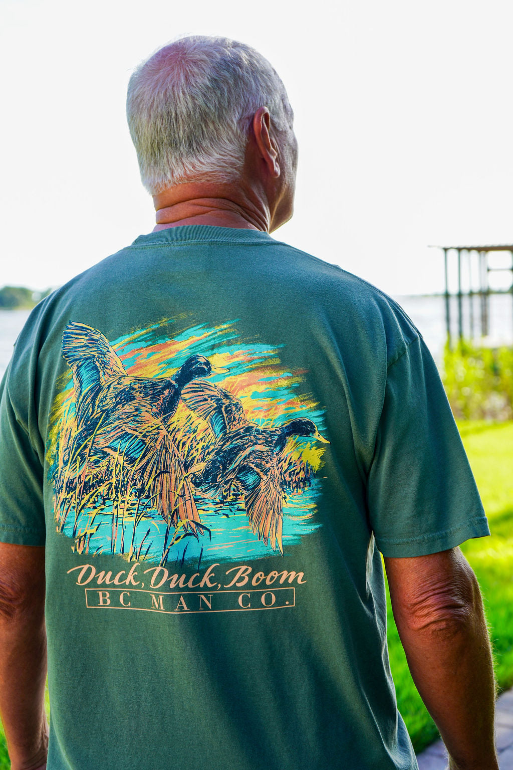 A man viewed from behind wearing a dark teal green t-shirt with a large graphic print of ducks on reeds and the text 'Duck Duck Boom BC MAN CO.' on the back, set against an outdoor background.